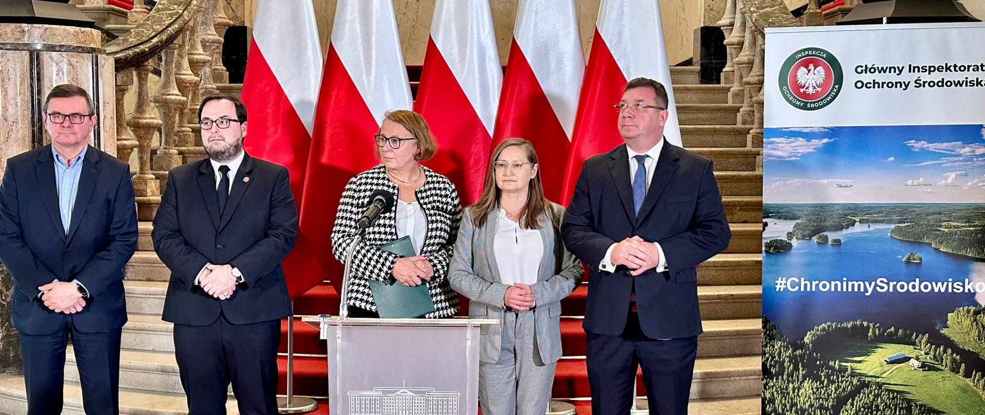 Pięć osób, dwie kobiety i trzech mężczyzn stoi za mównicą. Za nimi znajdują się polskie flagi, który przykrywają marmurowe schody na wyższe piętro.