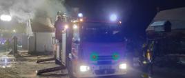 Na zdjęciu widać strażacką autodrabinę mechaniczną. Strażacy gaszą pożar poddasza z kosza drabiny znajdującej się nad dachem. W tle po lewej stronie widać samochód pożarniczy z rozłożonym masztem oświetleniowym
