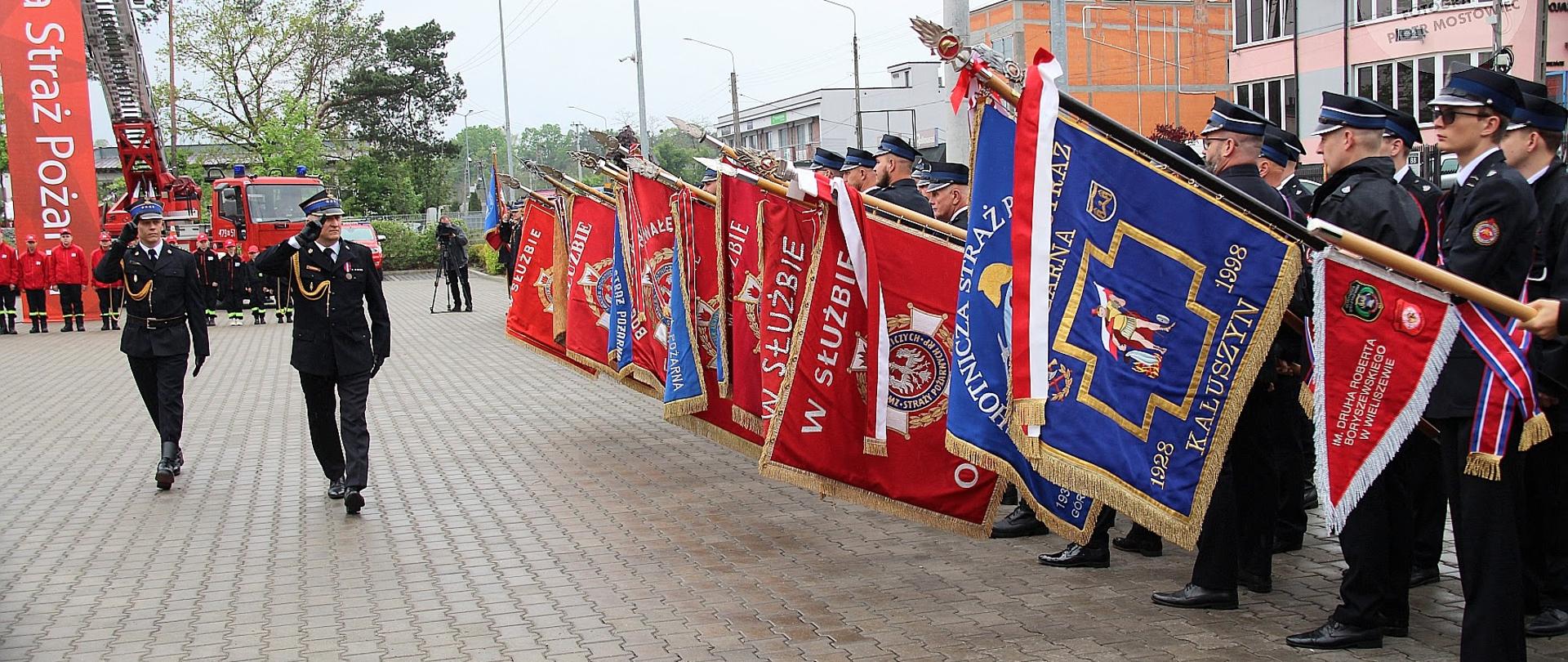 Fotografia przedstawia ceremonialny przemarsz uczestników uroczystości pomiędzy dwoma rzędami pocztów sztandarowych. Strażacy stoją w zwartej formacji, trzymając kolorowe sztandary i flagi jednostek strażackich, co podkreśla podniosły charakter wydarzenia. Całość odbywa się na utwardzonym placu przed budynkiem straży pożarnej.