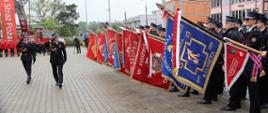 Fotografia przedstawia ceremonialny przemarsz uczestników uroczystości pomiędzy dwoma rzędami pocztów sztandarowych. Strażacy stoją w zwartej formacji, trzymając kolorowe sztandary i flagi jednostek strażackich, co podkreśla podniosły charakter wydarzenia. Całość odbywa się na utwardzonym placu przed budynkiem straży pożarnej.
