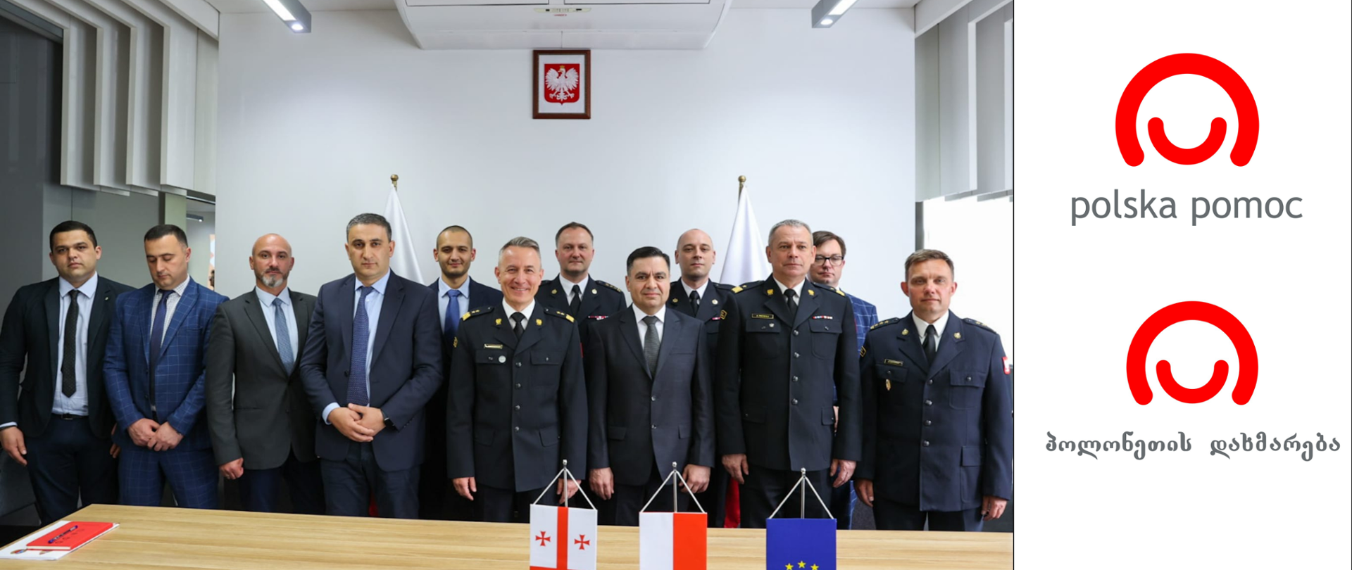 Grupa polskich strażaków oraz gruzińskiej delegacji stoją pozując do zdjęcia, przed nimi na stole flaga Polski, Gruzji i UE, na ścianie za grupą osób wisi portret orła białego