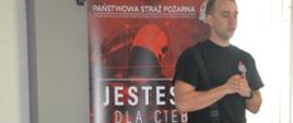 Szkolenie z pierwszej pomocy. Świetlica Komendy Powiatowej Policji w Rawiczu. Na tle roll-up promującego Państwową Straż Pożarną, na którym widnieje hasło "JESTESMY DLA CIEBIE", stoi strażak prowadzący szkolenie. 
