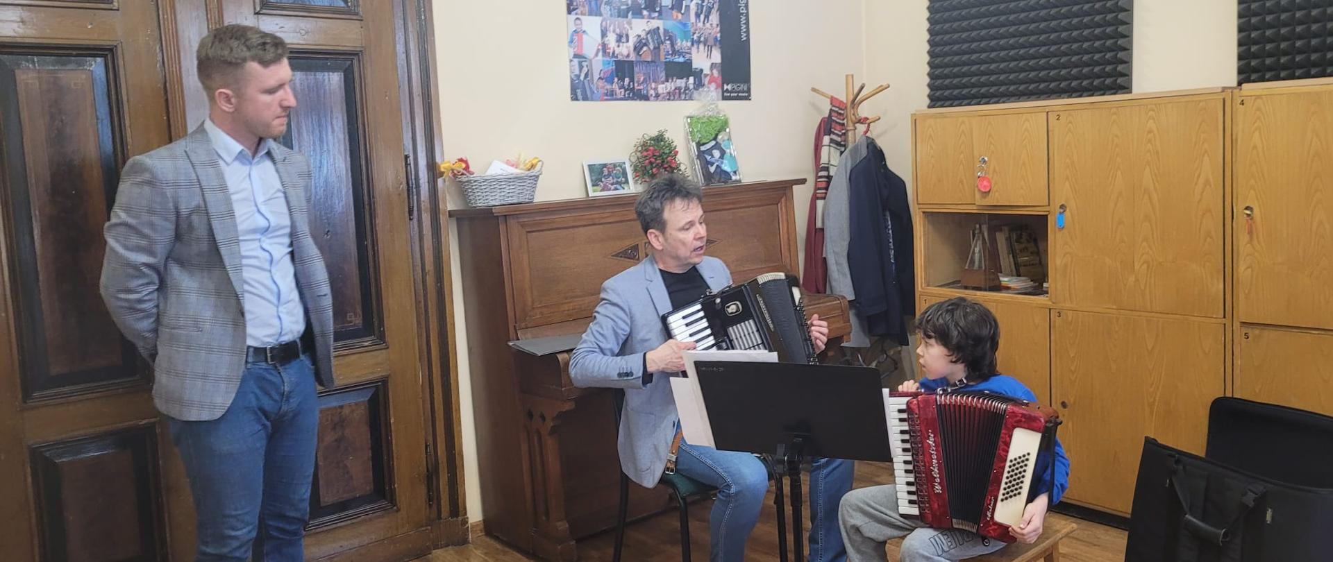 Od lewej stojący nauczyciel z klasy akordeonu P. T. Brusiński, który przygląda się jak prof. Piotr Chołołowicz udziela wskazówek gry na akordeonie uczniowi, oboje siędzą na krzesłach grając na instrumencie
