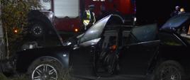Wypadek w Stwolnie. Na pierwszym planie stojący w rowie czarny samochód osobowy, który czołowo uderzył w drzewo. Auto ma uszkodzony przód. Otwarta jest pokrywa silnika oraz drzwi z lewej strony i bagażnik. W tle czerwony wóz strażacki, który na drzwiach ma naklejony numer 571P21. Pomiędzy samochodami stoi policjant w białej czapce oraz inny mężczyzna.