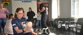 Szkolenie z pierwszej pomocy. Świetlica Komendy Powiatowej Policji w Rawiczu. Na pierwszym planie umundurowana funkcjonariuszka policji, która klęcząc na prawym kolanie, na lewym trzyma manekina "niemowlaka" - wykonując czynności ratujące życie po uprzednim zakrztuszeniu się dziecka. W tle strażak i 6 innych uczestniczek szkolenia. 