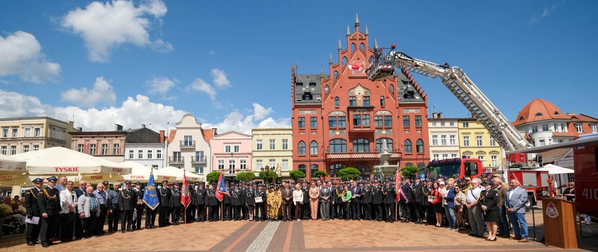 Uczestnicy strażackiej uroczystości pozują do wspólnego zdjęcia na rynku miejskim. Na pierwszym planie stoją strażacy w galowych mundurach, poczty sztandarowe i goście, a w tle znajduje się czerwony ratusz oraz wysięgnik strażacki z zawieszoną polską flagą.