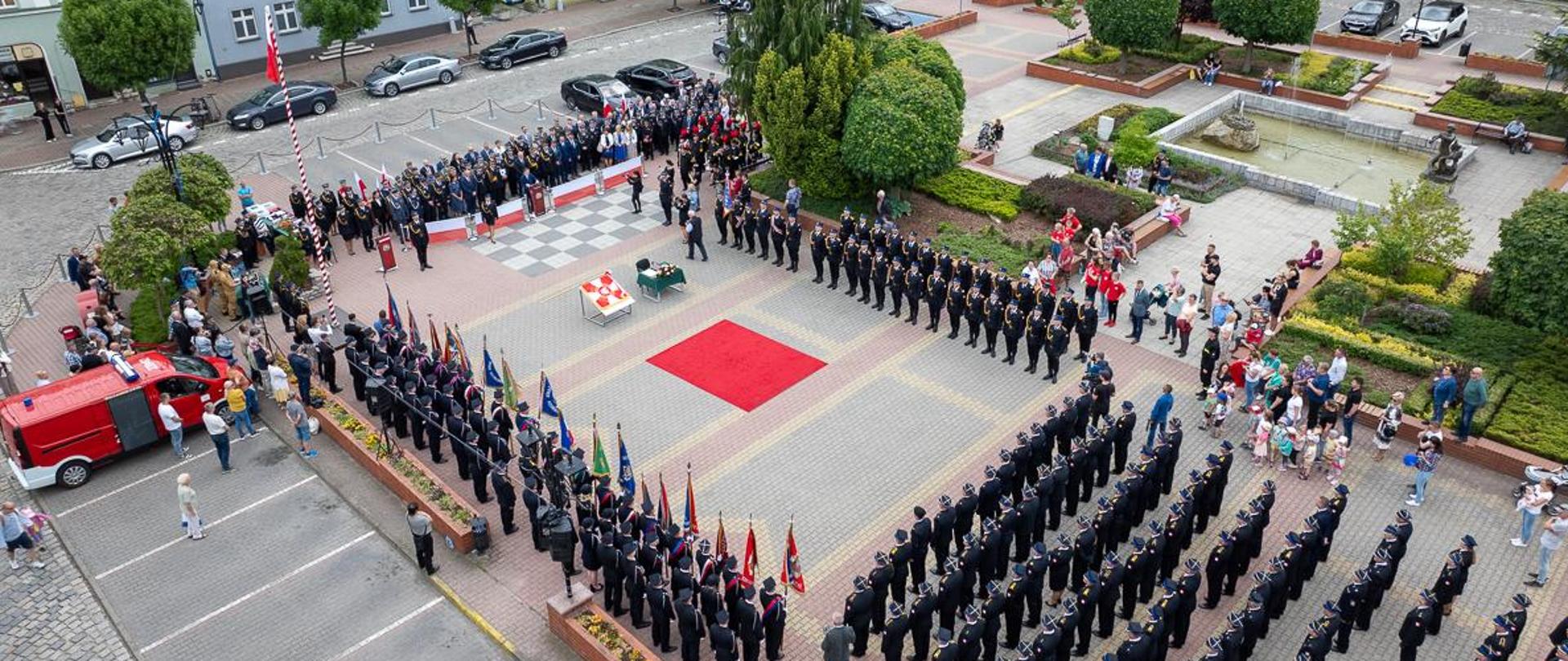 Ujęcie krapkowickiego rynku z drona podczas trwającej uroczystości z Okazji Wojewódzkich Obchodów Dnia Strażaka w Krapkowicach. Na środku widać czerwony dywan, a przy nim ułożony na specjalnym stojaku sztandar dla KP PSP Krapkowice. W lewej górnej części zdjęcia widać trybunę honorową z zaproszonymi gośćmi. W górnej części ustawiona kompania honorowa, a naprzeciwko pododdział OSP ze sztandarami. W prawym dolnym rogu pododdział odznaczonych i awansowanych. Wokół wydarzenia zgromadzeni mieszkańcy i sympatycy straży pożarnej. 