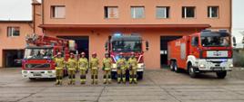 Na zdjęciu widać funkcjonariuszy PSP w Kaliszu, którzy stoją w szeregu przed garażami JRG 2 na ulicy Częstochowskiej. Funkcjonariusze mają piaskowy kolor mundurów i czerwone hełmy. Za plecami ratowników w szeregu stoją wyprowadzone z garażu samochody strażackie. Samochody są w kolorze czerwonym. 