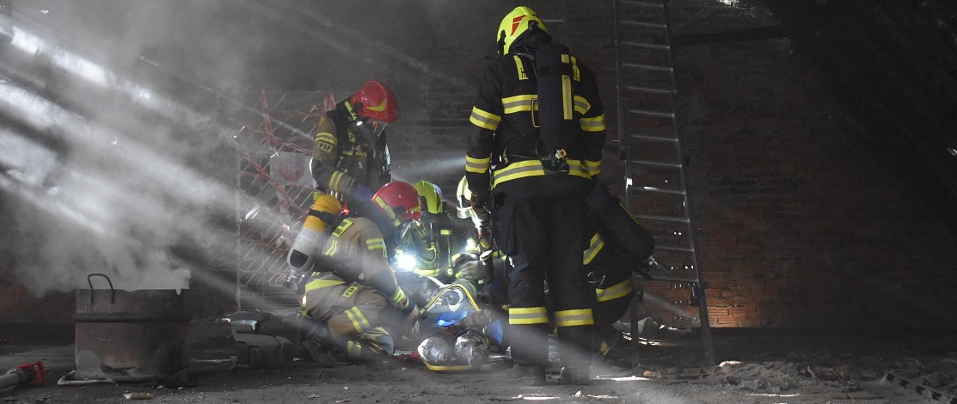 Grupa strażaków, w tym ratownicy z Czech udzielają pomocy osobie poszkodowane (manekin). Strażacy w aparatach ochrony dróg oddechowych. Miejsce działań na strychu.