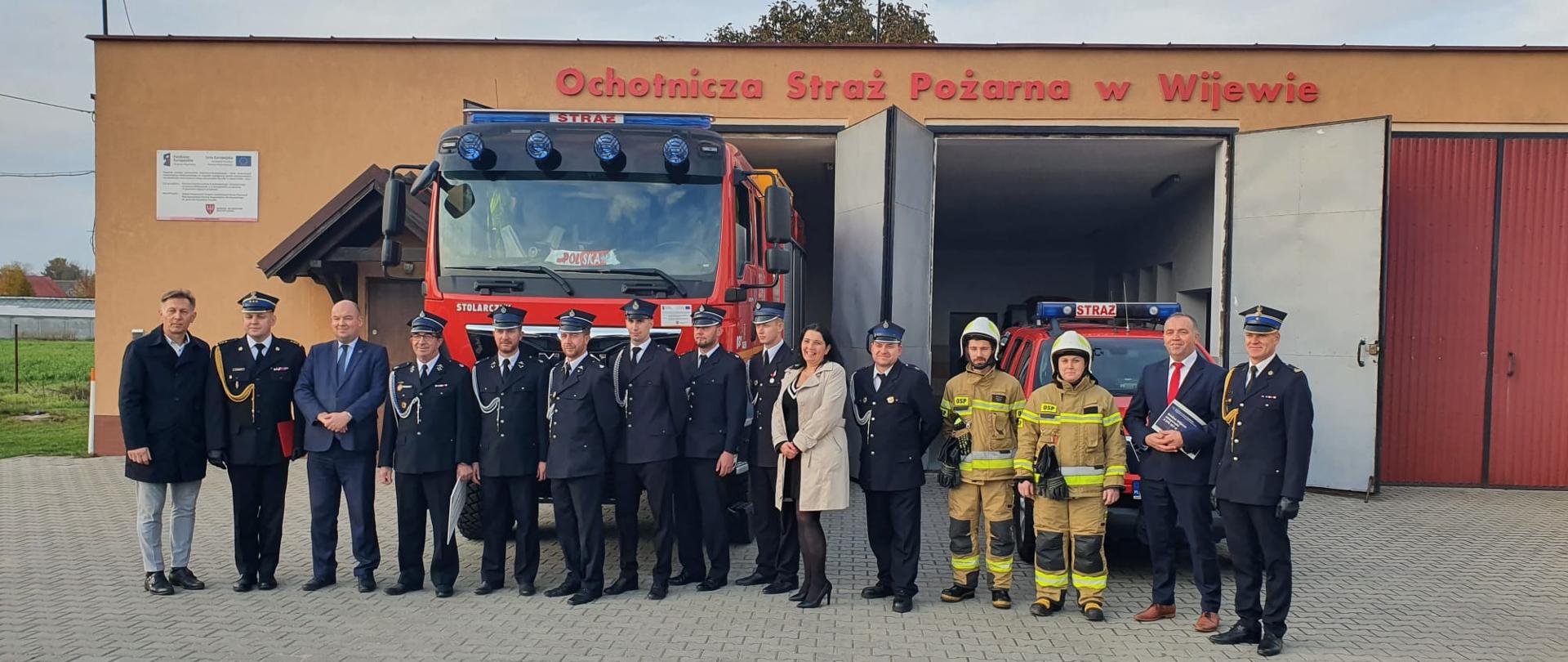 Strażacy ochotnicy i strażacy PSP stoją przed remizą. W tle samochody strażackie.