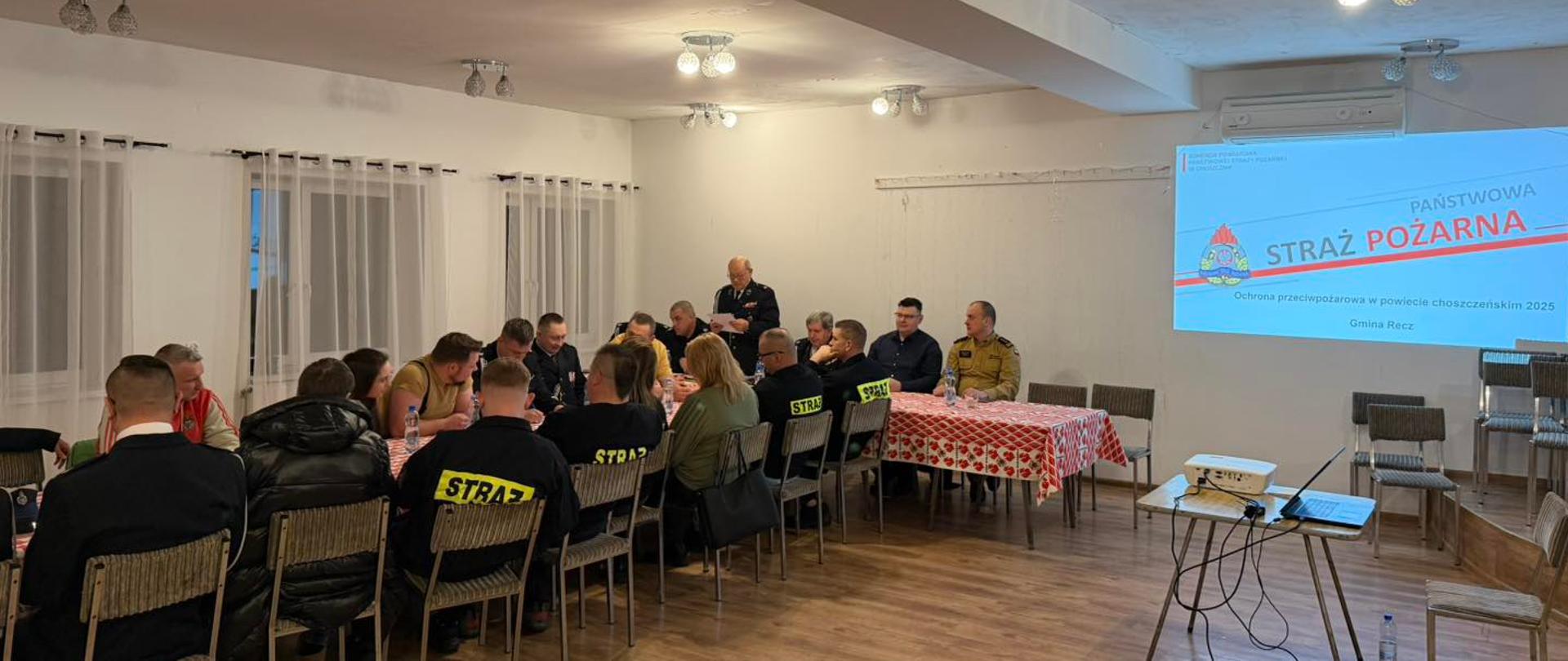 Uczestnicy zebrania siedzą przy długim stole w sali. Część osób ubrana jest w mundury strażackie i odzież służbową. W tle na ścianie widnieje prezentacja multimedialna.