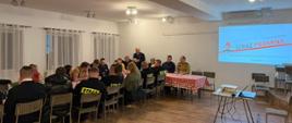 Uczestnicy zebrania siedzą przy długim stole w sali. Część osób ubrana jest w mundury strażackie i odzież służbową. W tle na ścianie widnieje prezentacja multimedialna.