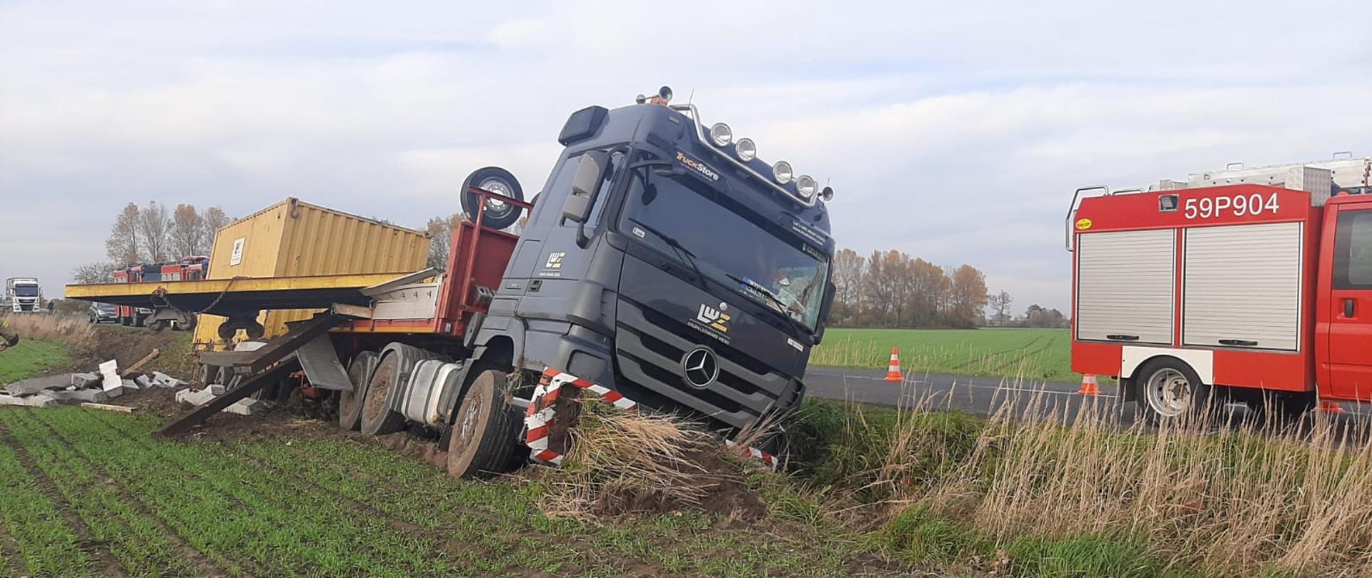 Widać samochód ciężarowy z przyczepą w rowie, po prawej samochód strażacki