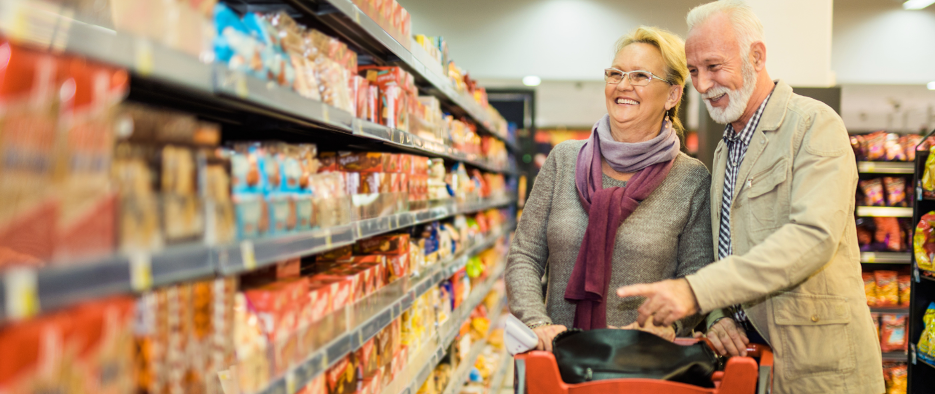 Uśmiechnięci seniorzy wybierają produkty z półek w alejce sklepowej.