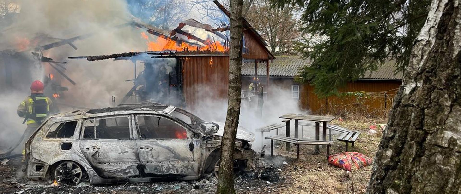 Przed palącym się domkiem letniskowym stoi spalony samochód osobowy. Strażacy gaszą pożar. 