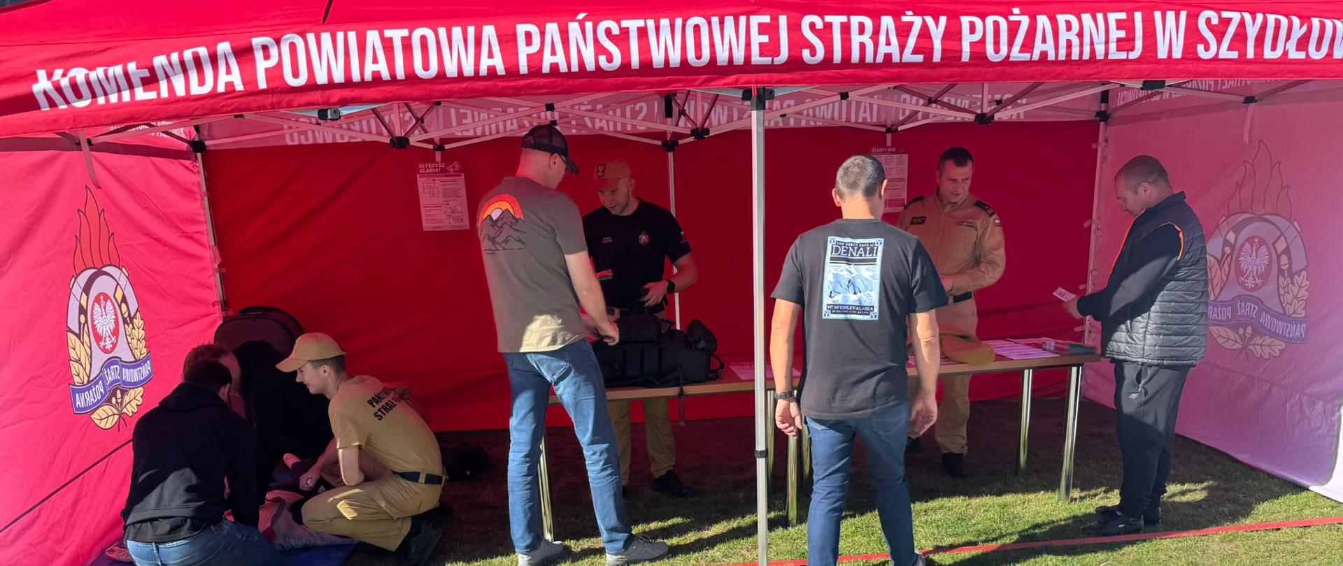 Grupa osób i strażaków przebywa pod dużym, czerwonym namiotem promocyjnym z białym napisem „Komenda Powiatowa Państwowej Straży Pożarnej w Szydłowcu”. Po lewej stronie na macie odbywa się pokaz pierwszej pomocy, podczas gdy inni mężczyźni rozmawiają ze strażakami przy stole w głębi namiotu.