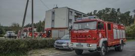 Przed budynkiem objętym działaniami widać podnośnik hydrauliczny oraz dwa samochody gaśnicze. Dodatkowo przy pojazdach straży pożarnej zaparkowany został radiowóz Policyjny. W oddali widać las.Jeden z pojazdów gaśniczych stoi na drodze blokując jeden pas. 