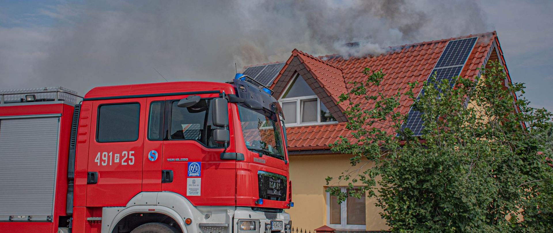 ZDJĘCIE PRZEDSTAWIA SAMOCHÓD POŻARNICZY, ZA NIM BUDYNEK, W KTÓRYM DOSZŁO DO POŻARU. WIDOCZNY DYM WYDOBYWAJĄCY SIĘ SZCZYTEM DACHU