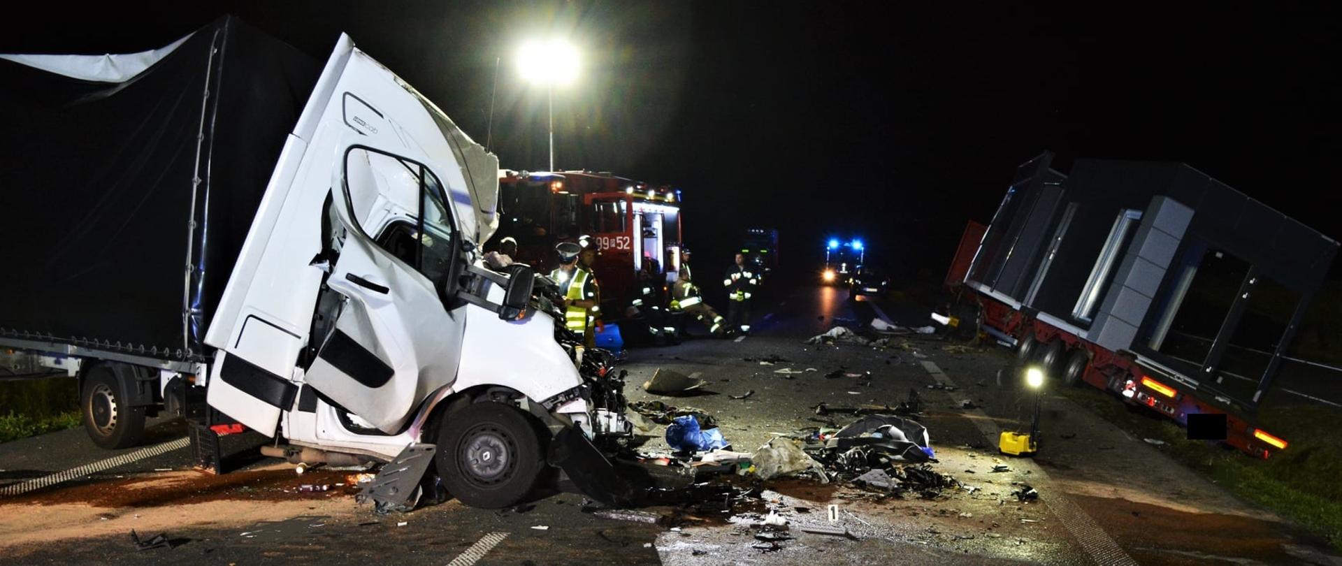 Po lewej widać uszkodzony przód samochodu dostawczego, po prawej samochód ciężarowy w rowie, w oddali samochód straży pożarnej, jest noc
