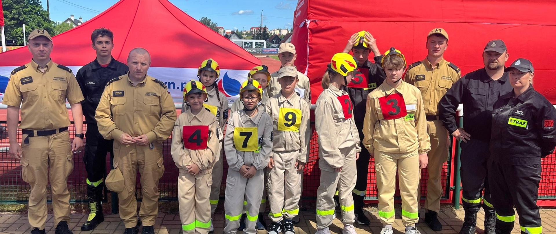 Na zdjęciu pozuje grupa Młodzieżowej Drużyny Pożarniczej z nimi kilku strażaków. W tle czerwone namioty.