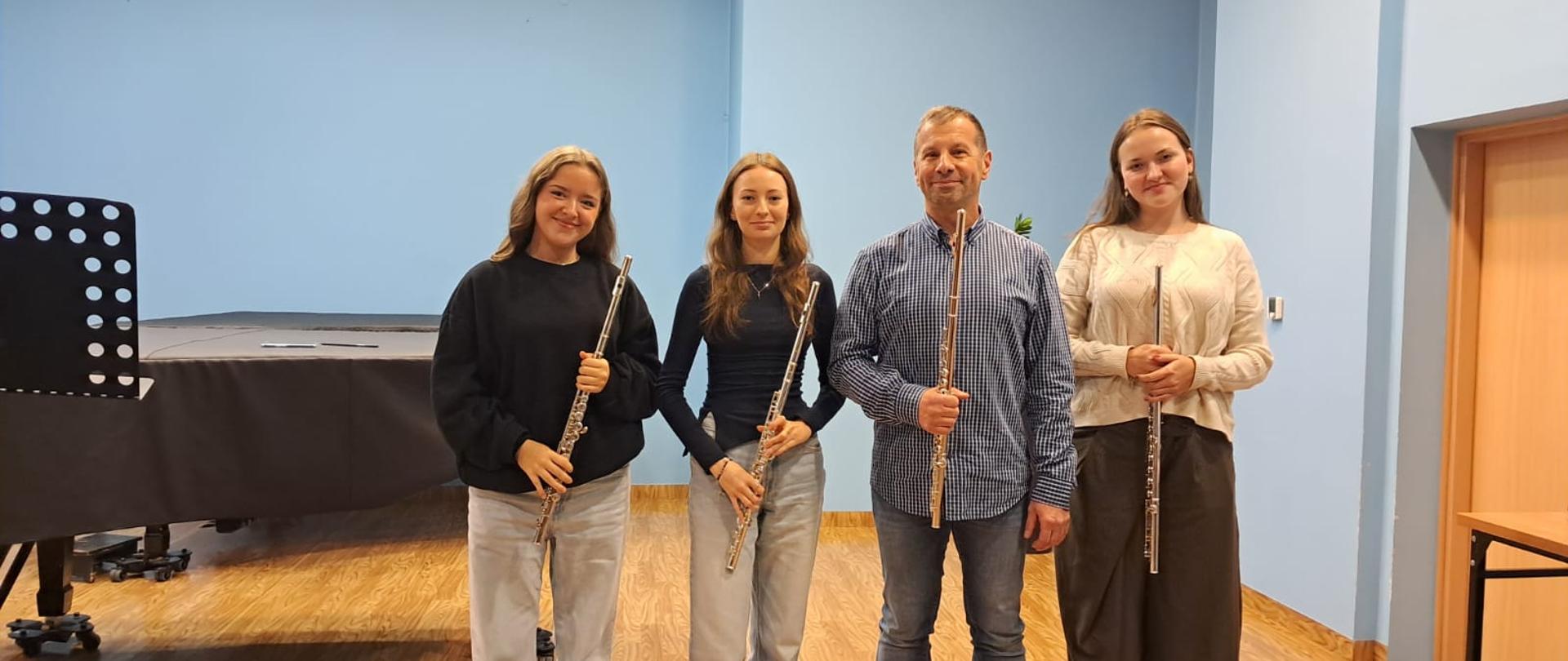 Kolorowe zdjęcie, na którym znajdują się 4 osoby stojące w jednym rzędzie (3 uczennice i dr hab. Łukasz Ziemnik). Wszystkie osoby trzymają w rękach srebrne flety. Za nimi fortepian i niebieska ściana.