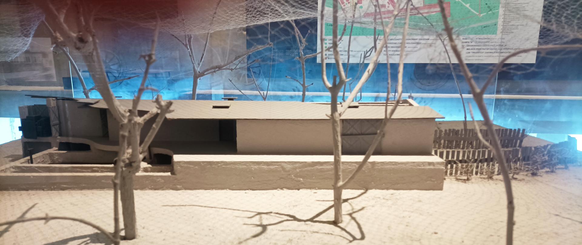 Makieta komory śmierci – na zdjęciu widzimy makietę komory gazowej, która znajduje się w Muzeum- Miejsce Pamięci w Bełżcu. Przed makietą ustawione są suche drzewa a nad rozciągnięta jest siatka maskująca.