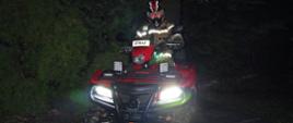 Widok z przodu. Pora nocna - strażak w ubraniu specjalnym i kasku ochronnym siedzi na quadzie.