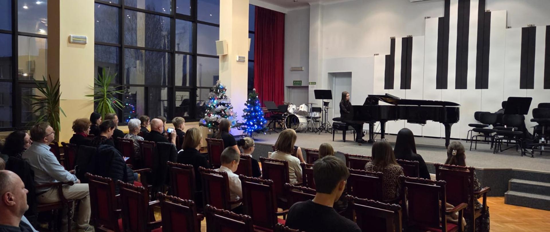 Publiczność na widowni, w tle widać dziewczynę grającą na fortepianie.