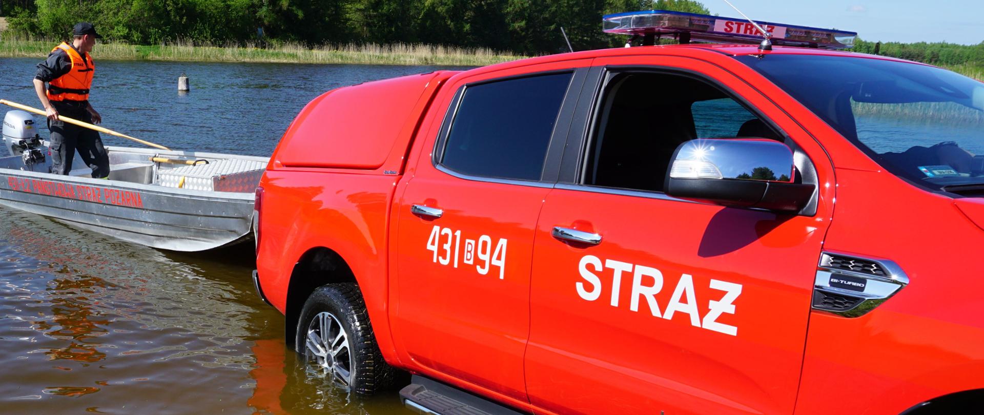 Czerwony strażacki samochód wjechał do wody z przyczepką na której jest łódź płaskodenna w celu zwodowania łodzi. Na łodzi stoi strażak w kamizelce ratunkowej.
