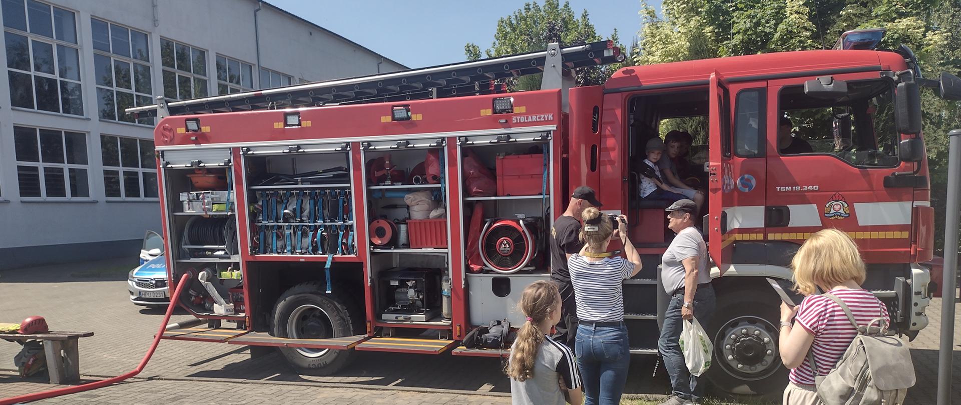 Zdjęcie w ciągu dnia. Duże nasłonecznienie. Widać samochód ratowniczo-gaśniczy, dzieci oraz funkcjonariuszy PSP.