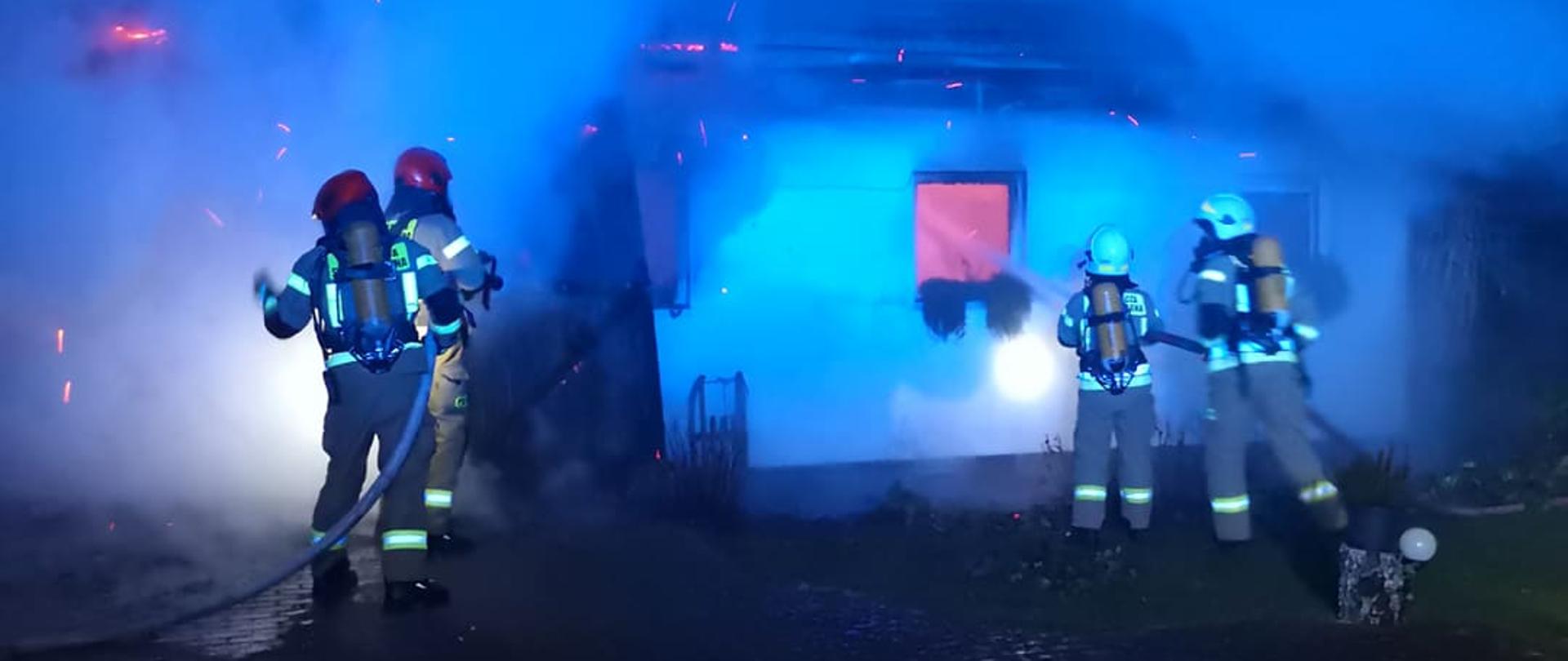 Zdjęcie przedstawia pożar budynku gospodarczego, wydobywa się z niego ogień i dym. Przed budynkiem stoją dwie roty strażaków - dwóch po lewej stronie zdjęcia (w czerwonych hełmach) i dwóch po prawej (W białych hełmach). Trzymają oni w rękach prądownice wodne wraz z wężami. Strażacy są ubraniu w ubranie specjalne, hełmy i sprzęt ochrony układu oddechowego. Zdjęcie wykonano nocą.