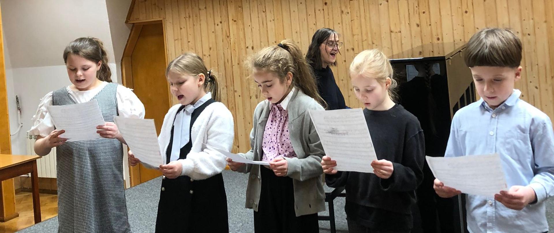 Dzieci stojąc na scenie śpiewają z nut, a za nimi kobieta akompaniuje na fortepianie.