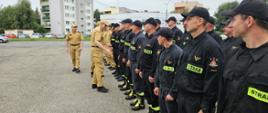 Strażacy podczas przywitania z Zastępcą Komendanta Głównego oraz Komendantem Wojewódzkim. Ustawieni w dwuszeregu stoją na placu ubrani w piaskowe mundury oraz czarne ubrania koszarowe. Komendanci witają się z załogą poprzez podanie ręki z tyłu widoczne budynki oraz samochody