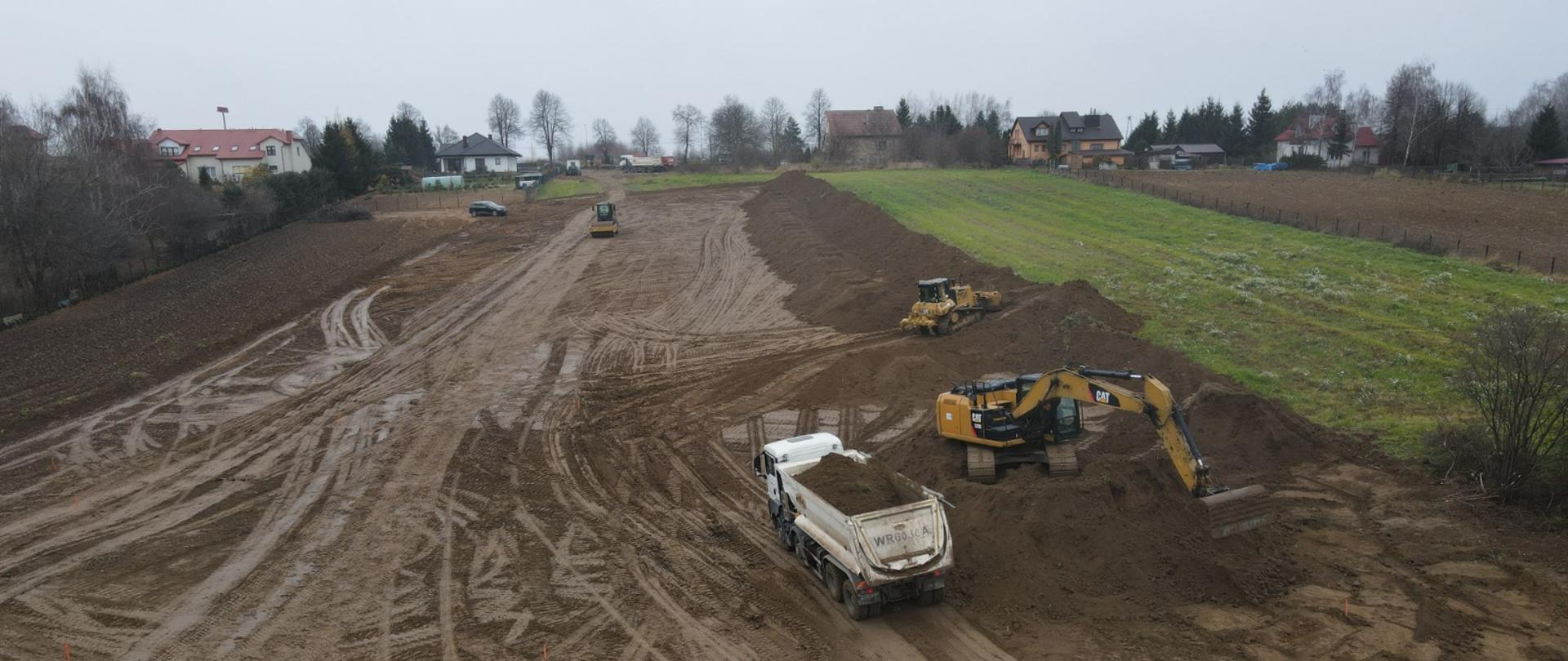 DK9 obwodnica Ostrowca Świętokrzyskiego - roboty ziemne, na pasie ziemi koparki, walce, ciężarówki, w oddali domy, obok pole uprawne 