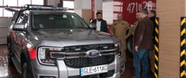 W garażu komendy obok szarego samochodu specjalnego rozpoznawczo-ratowniczego Ford Ranger Limited stoją i rozmawiają Starosta Lęborski, Komendant Powiatowy Państwowej Straży Pożarnej w Lęborku, Dowódca zmiany w komendzie, Zastępca Komendanta Powiatowego Państwowej Straży Pożarnej, kierownik Referatu Bezpieczeństwa i Zarządzania Kryzysowego Starostwa Powiatowego w Lęborku. Na boku pojazdu widoczny czerwony pas odblaskowy z napisem straż. Na dachu pojazdu belka ostrzegawcza straż. Za osobami stoi czerwony ciężki samochód ratowniczo - gaśniczy.