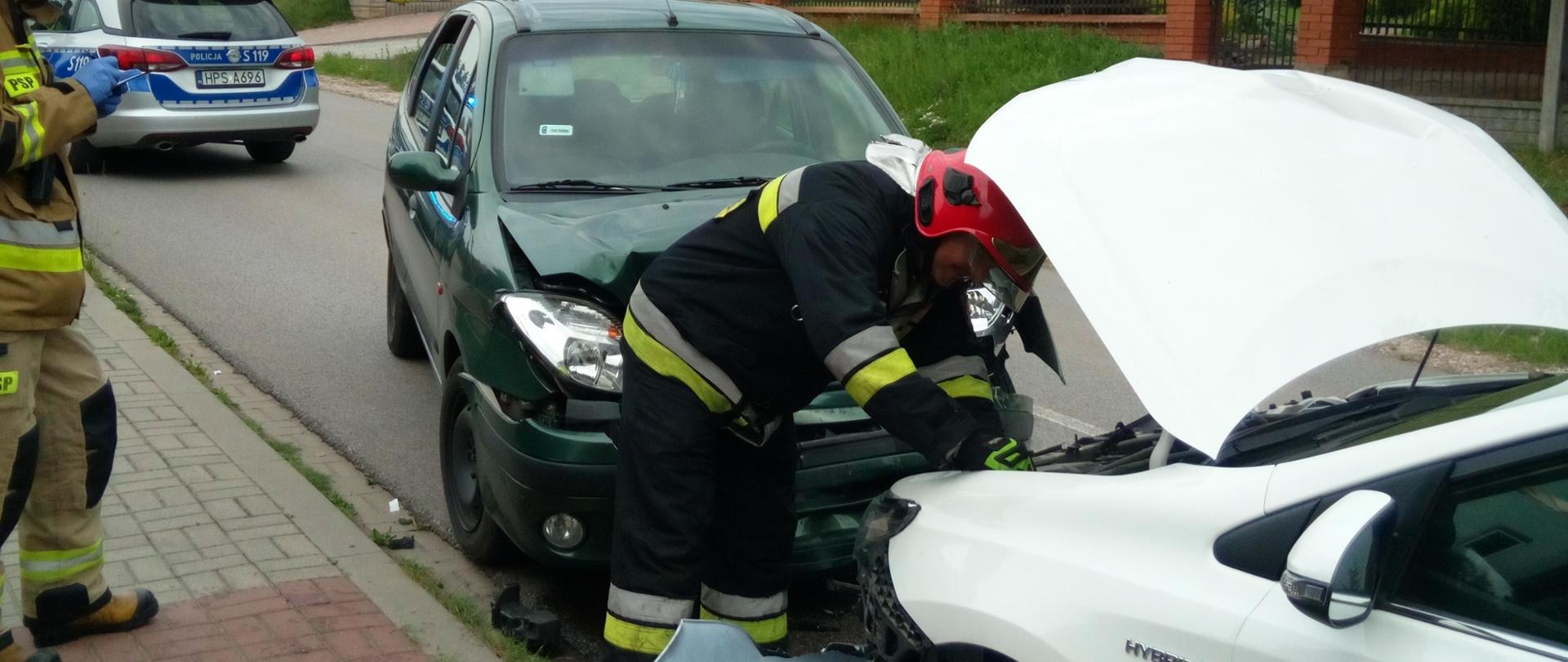 Fotografia przedstawia dwa samochody osobowe, które uległy zderzeniu. W dalszej perspektywie widać radiowóz. Przy uszkodzonych autach stoją strażacy.