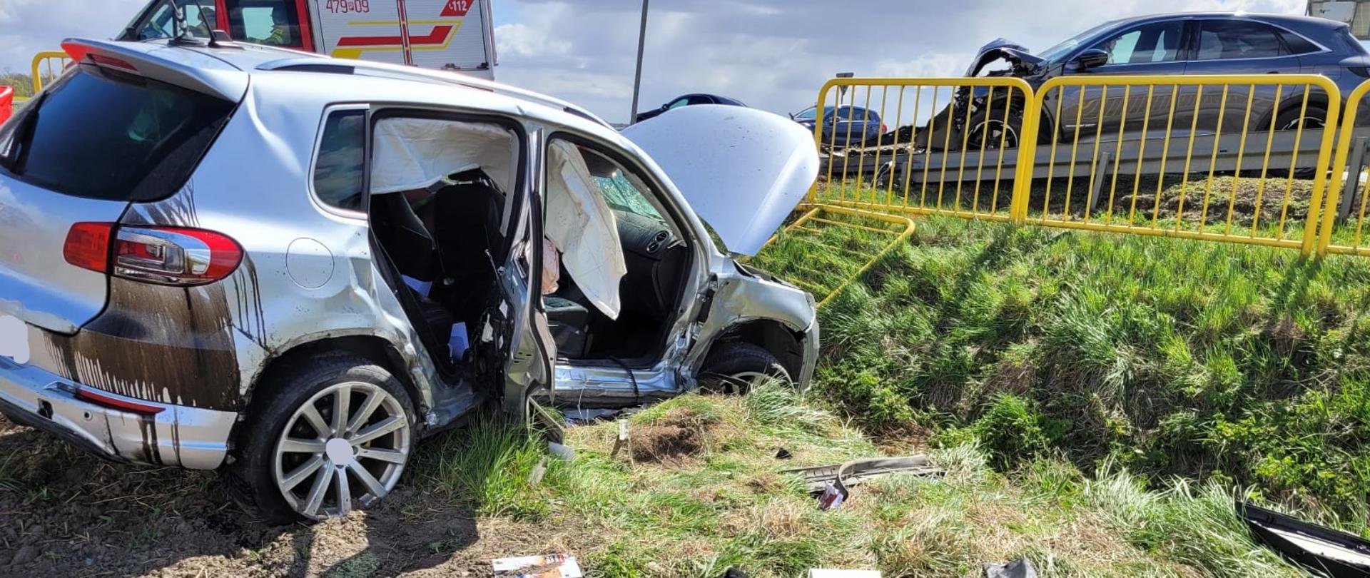 Rozbity biały samochód osobowy stoi w rowie przy drodze, z poważnie uszkodzonym bokiem i otwartymi drzwiami. Wewnątrz widać wystrzelone poduszki powietrzne. W tle znajduje się wóz strażacki oraz drugi uszkodzony samochód na jezdni, oddzielony żółtą barierką. Na ziemi wokół auta leżą fragmenty karoserii i przedmioty z wnętrza pojazdu.