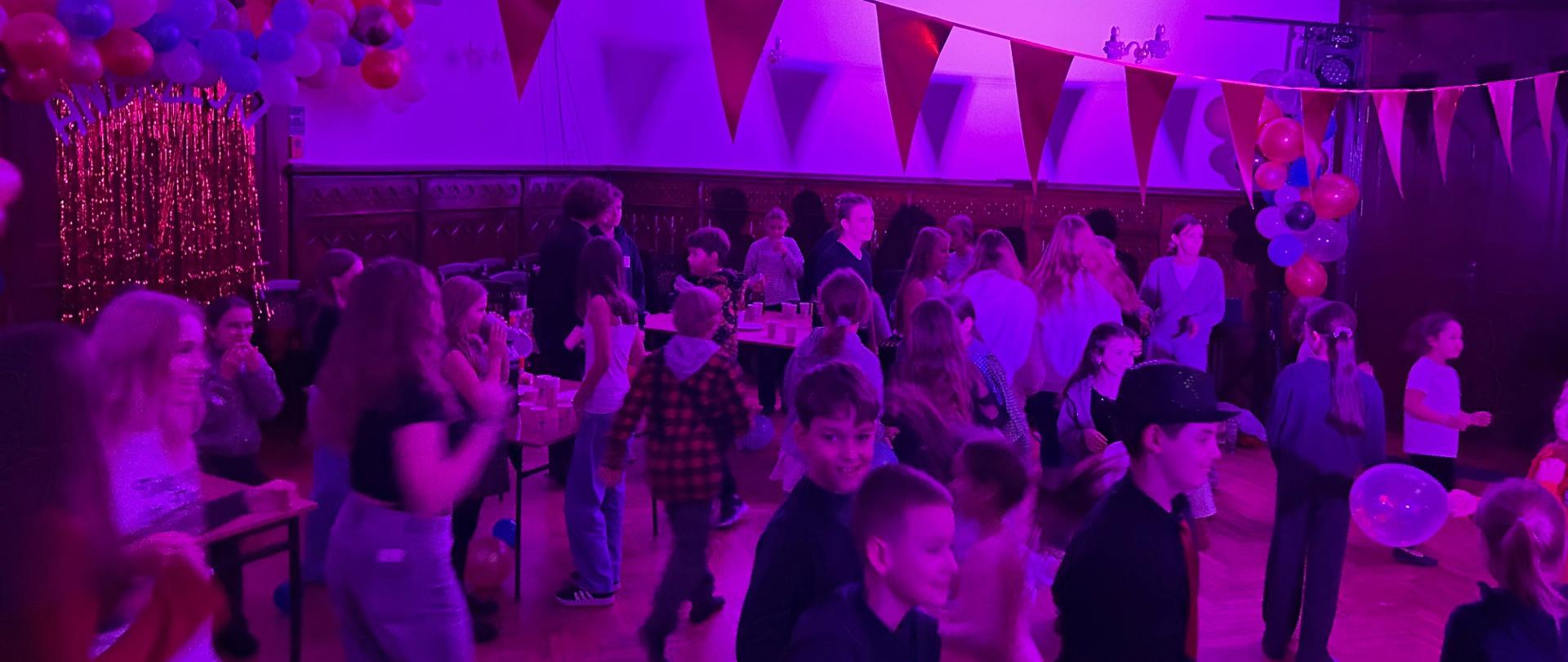 Grupa uczniów bawiących się i tańczących w udekorowanej sali podczas szkolnych andrzejek, oświetlonej fioletowym światłem i ozdobionej balonami oraz girlandami.