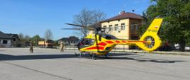 Zdjęcie przedstawia stojący na placu żółty śmigłowiec Lotniczego Pogotowia Ratunkowego. Przed śmigłowcem stoi dwóch strażaków ubranych w piaskowe ubrania specjalne na głowie maja hełmy. W oddali widać samochód ciężarowy i żółty budynek.