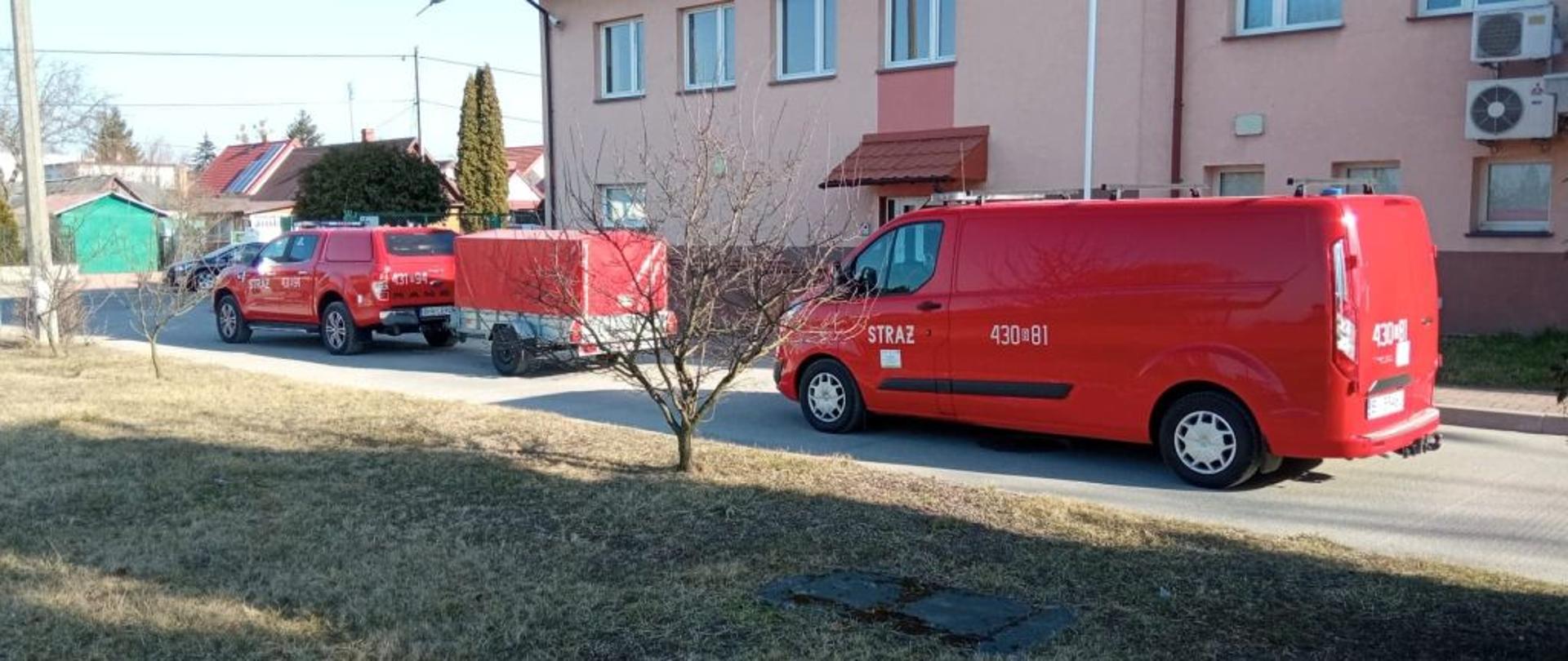 Dwa czerwone osobowe samochody strażackie. Pierwszy ciągnie przyczepkę z czerwoną plandeką.