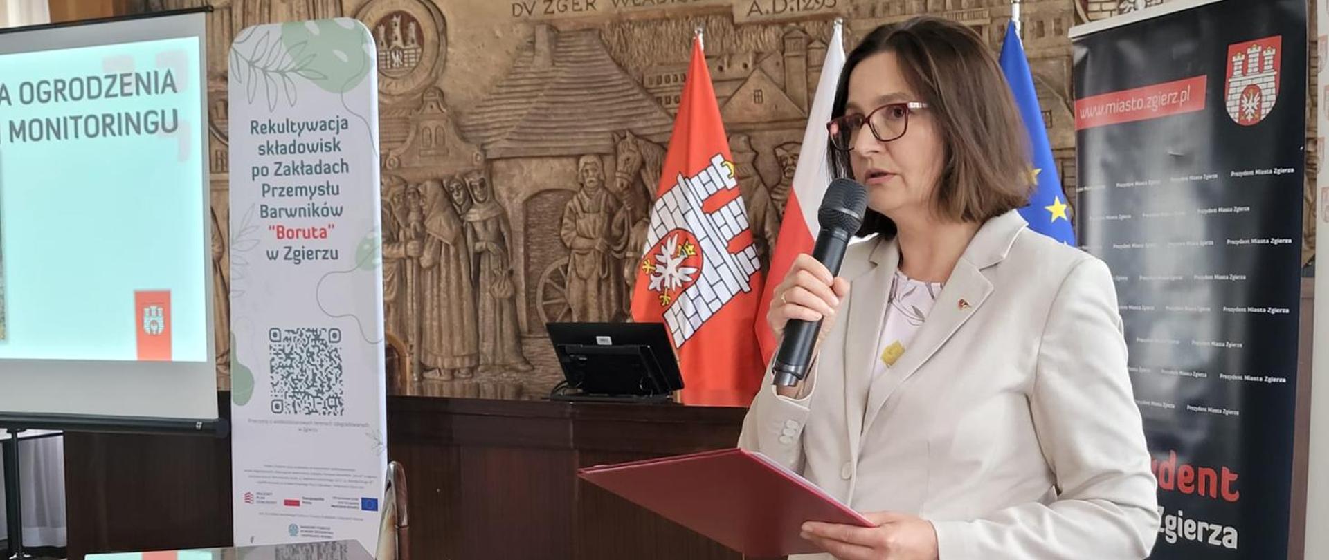 Deputy Minister of Climate and Environment Anita Sowińska during a meeting reviewing the results of the work carried out to date by the Zgierz Task Force for Large-Scale Degraded Areas. The photograph shows the Deputy Minister speaking into a microphone while holding a burgundy folder in her hand.