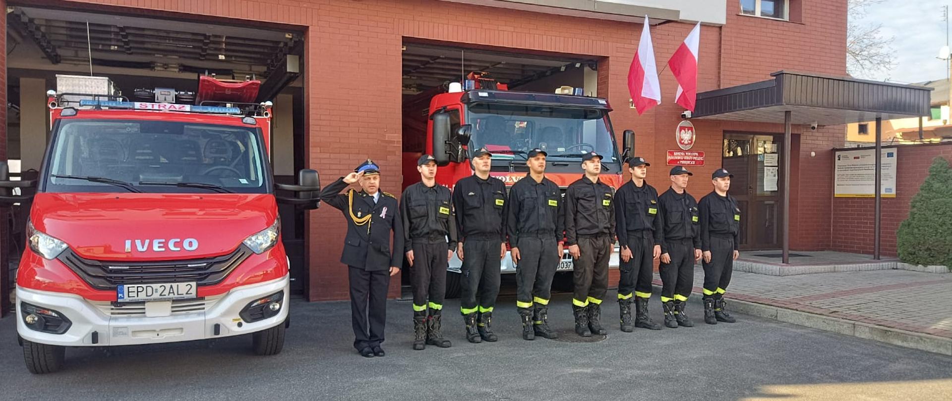 przed budynkiem komendy w jednym szeregu stoi 8 strażaków na baczność. Jeden z nich salutuje. Bramy wjazdowe do garaży są otwarte. Z tyłu widać 2 wozy strażackie 