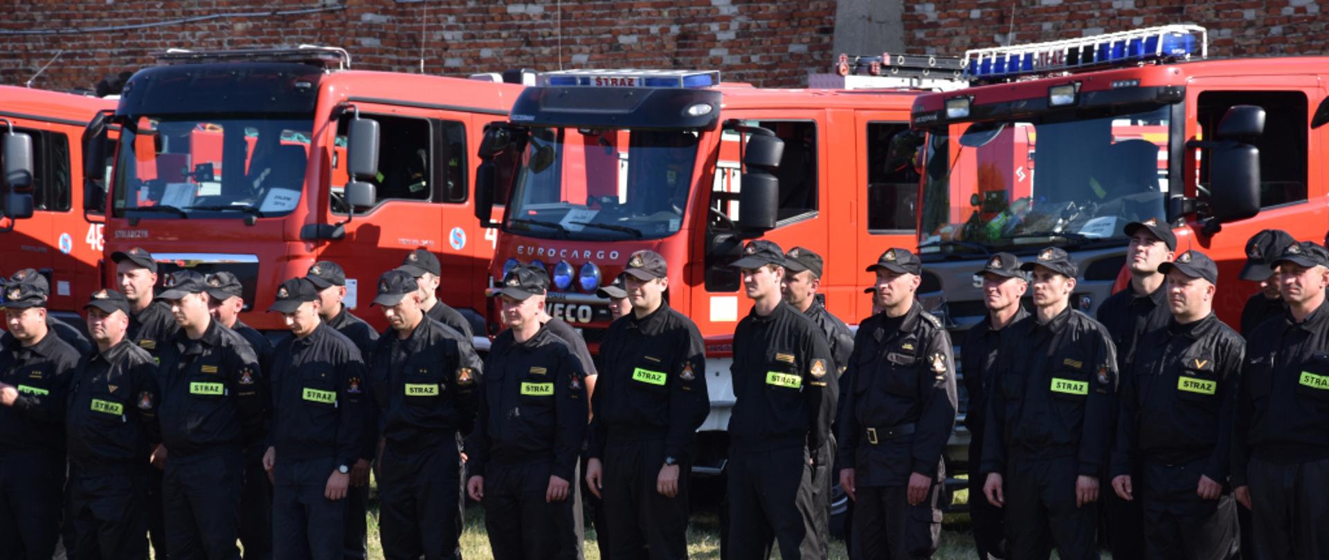 Na zdjęciu zbiórka strażaków, z tyłu za nimi stoją samochody pożarnicze