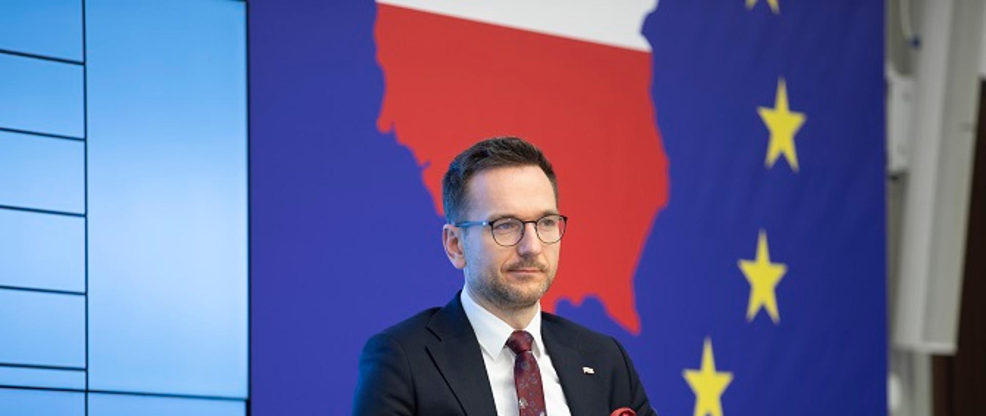 Minister Waldemar Buda siedzi na fotelu z dokumentami w ręku. Za nim duży ekran, na którym biało-czerwoną mapę Polski na granatowym tle z gwiazdkami, nawiązujące do flagi UE