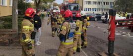 Strażacy w ubraniach specjalnych i hełmach stoją na osiedlowym chodniku podczas ćwiczeń. W tle znajdują się wozy strażackie i bloki mieszkalne.