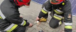 Dwóch strażaków miernikiem elektrycznym bada napięcie na świecącej żarówce podłączonej do paneli fotowoltaicznych.