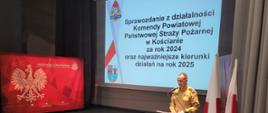 Strażacy i zaproszeni goście zebrani na sali podczas odprawy służbowej. Funkcjonariusz przemawia, w tle ekran z prezentowanym materiałem. 
