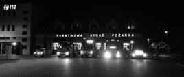 Czarno-białe zdjęcie. Przed budynkiem Komendy Powiatowej Państwowej Straży Pożarnej w Świeciu stoi 5 samochodów strażackich z włączonymi światłami alarmowymi pomiędzy nimi stoją strażacy. 