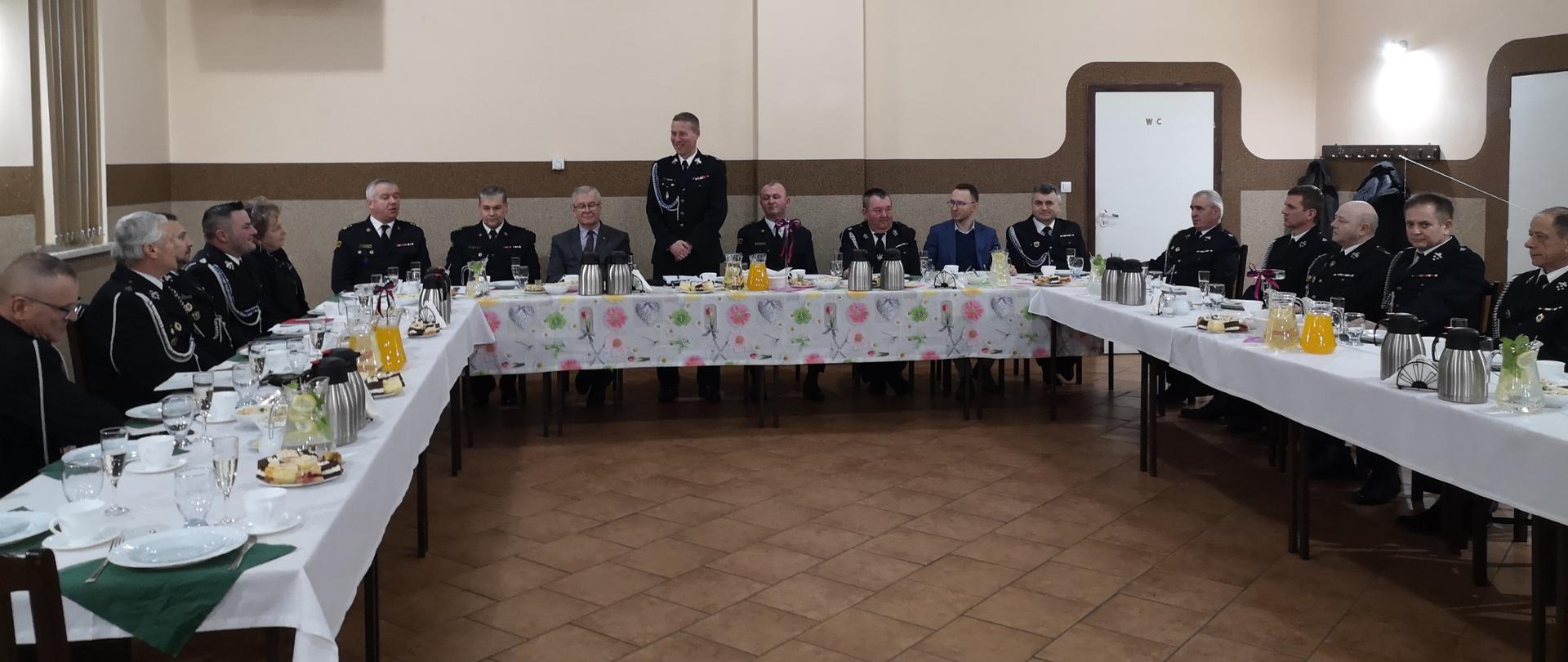 Na zdjęciu strażacy w ubraniach wyjściowych siedzą przy stole. Jeden z strażaków stoi na środku w trakcie przemowy. Zdjęcie wykonane w budynku remizy.