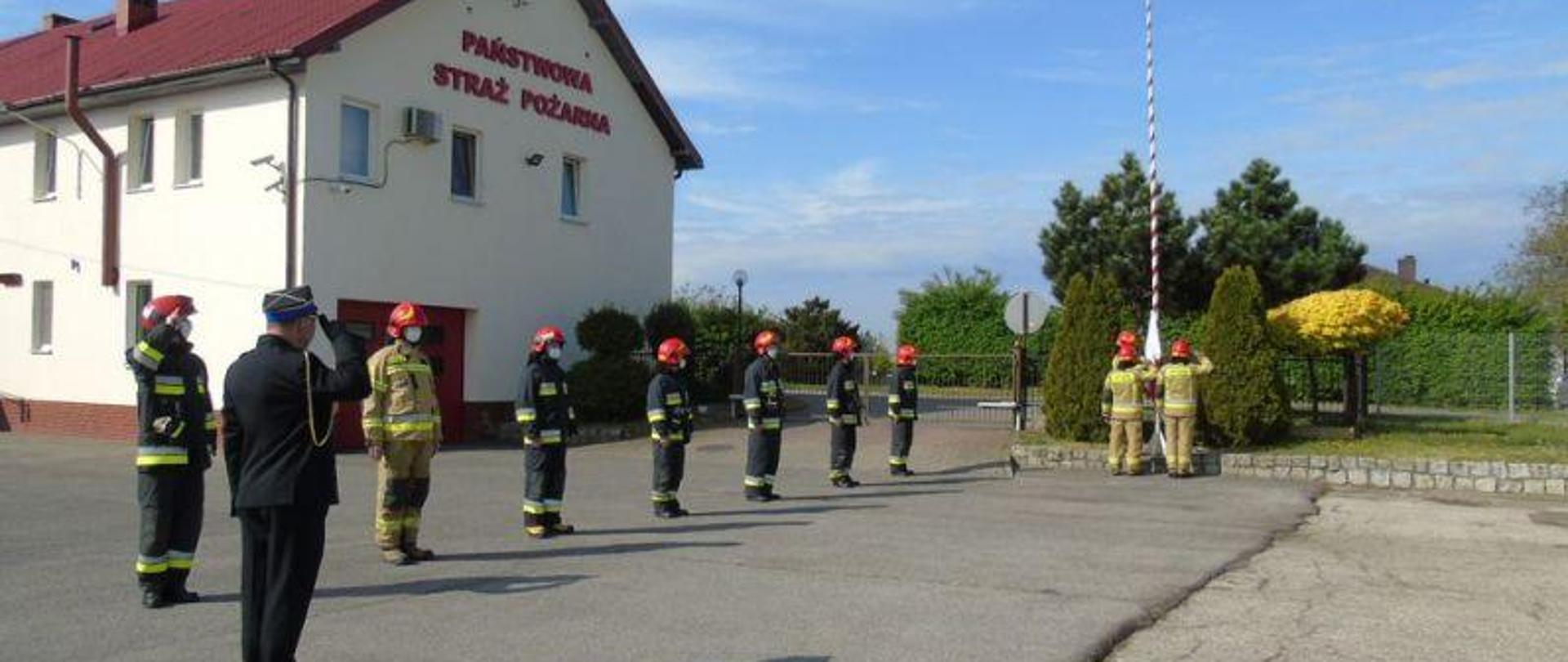 Zdjęcie strażaków w umundurowaniu w trakcie podnoszenia flagi państwowej na maszt.